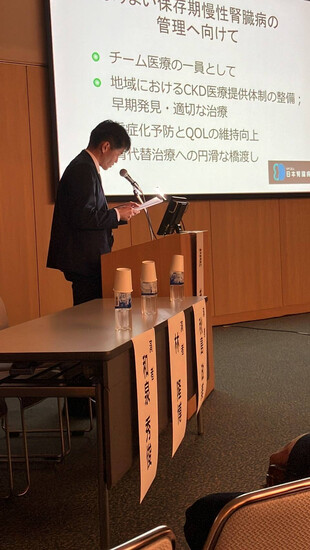 【画像】腎不全看護特定認定看護師 和泉秀俊『腎不全看護特定認定看護師として、患者さんの「これから」に寄り添うケアを』私は腎不全看護特定認定看護師として、慢性腎臓病（CKD）の早期から透析導入、さらには終末期に至るまで、患者さんの人生に寄り添う看護を実践しています。腎不全は長い経過をたどる疾患であり、治療選択や生活の変化に伴う不安、葛藤を抱える方も少なくありません。だからこそ、医療的な支援だけでなく、心のケアや意思決定支援がとても大切だと感じています。近年では「慢性腎臓病透析予防加算」の導入により、看護師が果たす役割も広がっています。私は、最新の診療ガイドラインをもとに、患者さんの自己管理支援や生活指導を行いながら、医師・薬剤師・管理栄養士・公認心理師・臨床工学技士・理学療法士・ソーシャルワーカー・看護補助者（当院呼称ケアメイト）など多職種と連携し、チームでの包括的なケアを推進しています。また、腎不全における緩和ケアの視点も、私の看護において欠かせない要素です。透析を選ばないという選択肢も含め、患者さんやご家族が納得して意思決定できるよう、対話を重ねながら支援しています。症状緩和や生活の質の維持を目指し、「その人らしく生きる」ことを支える看護を心がけています。院内では、多職種による勉強会やカンファレンスを定期的に開催し、CKDケアに関する最新の知見や実践を共有しています。さまざまな職種の視点を持ち寄ることで、より柔軟で患者さん本位の支援が可能になると実感しています。ちなみに私は、プロ野球・東京ヤクルトスワローズのファンでもあります。中でも、昨年引退した川端慎吾選手の大ファンでした。度重なるケガを乗り越え、代打の切り札としてチームに貢献し続けた姿には、何度も勇気をもらいました。どんな状況でも自分の役割を見つけ、全力で果たすその生き方に、私自身も強く憧れています。これからも、患者さんとチームの架け橋として、腎不全看護の専門性と緩和ケアの視点を活かしながら、日々の看護に真摯に向き合っていきたいと思います。